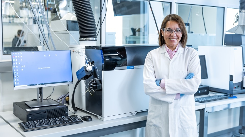 A technician in a white lab coat and glasses stands confidently in a bright laboratory beside sophisticated scientific equipment and a computer.