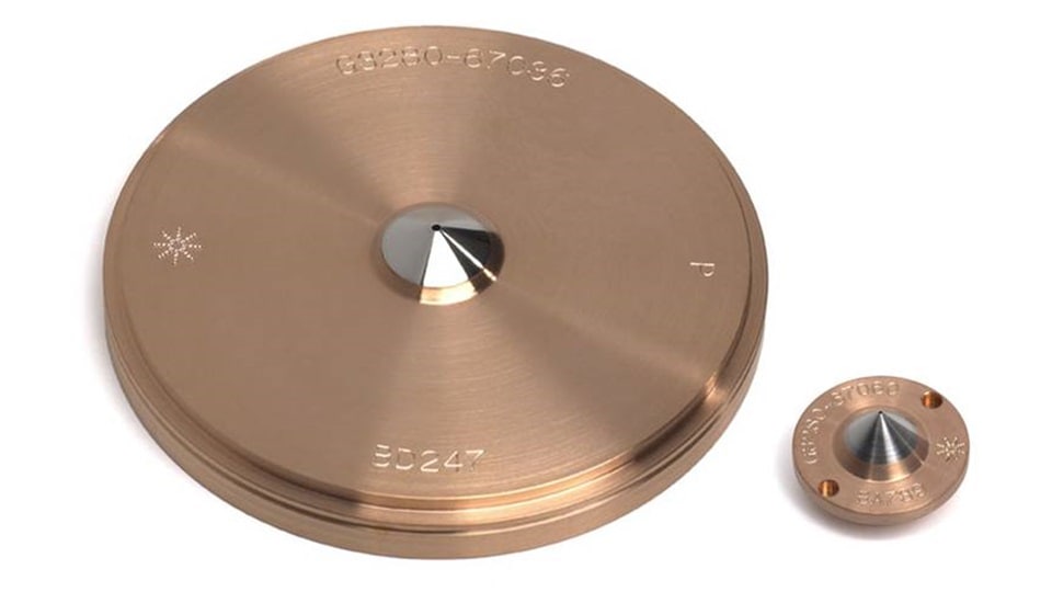 Copper-based conical sampling and skimmer cones with a platinum tip and engraved text, casting a shadow on a white surface. The metallic, polished finish gives a sense of precision engineering.