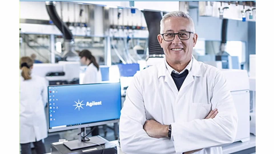 A smiling scientist in a lab coat stands confidently with arms crossed in a modern laboratory, surrounded by equipment and colleagues in lab attire.